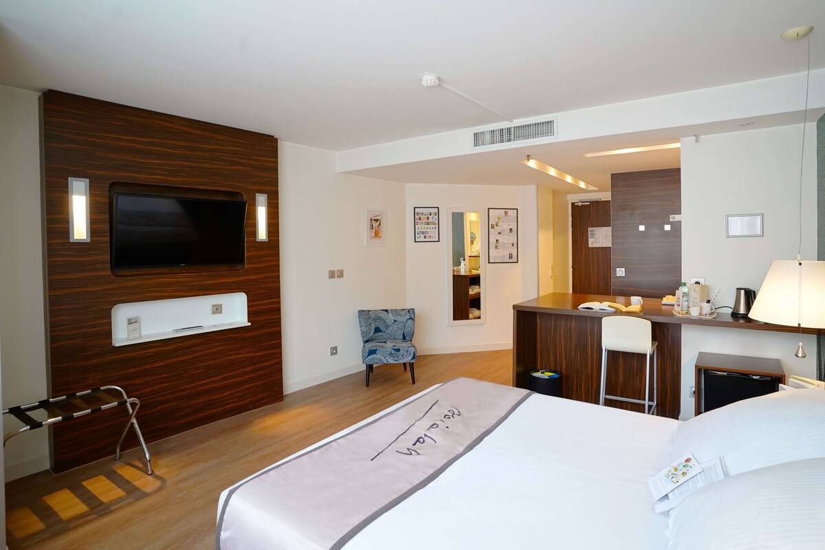 Habitación de hotel con cama, escritorio con lámpara, silla, televisor y espejo.