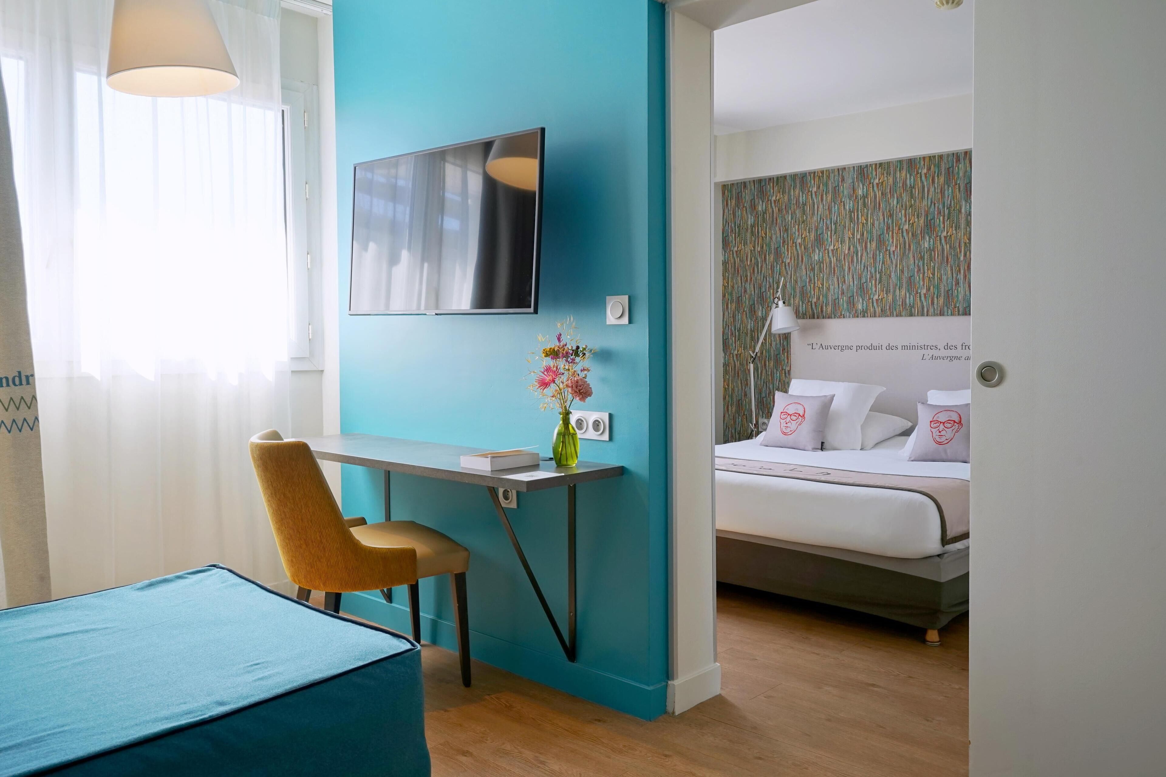Een moderne hotelkamer met een wand in turquoise, een bed met wit beddengoed, een bureau met stoel, en een vaas met bloemen.