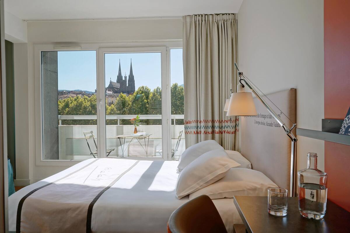 Una habitación de hotel con una cama, una mesa con un vaso y una botella, una lámpara, y vista a una iglesia por la ventana.