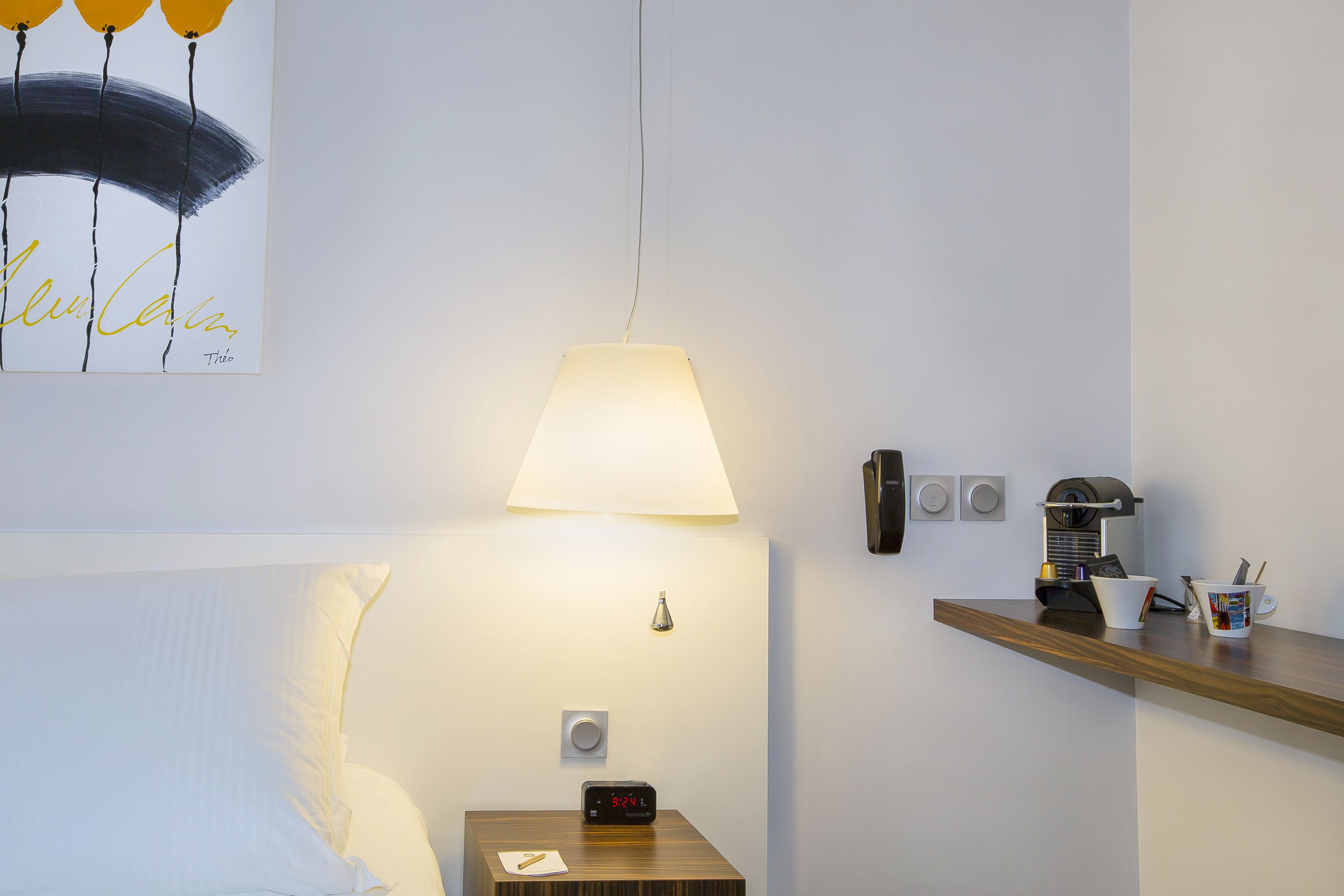 A hotel room with a bed, a nightstand with a clock, a lamp, a shelf with a coffee maker, and a painting on the wall.