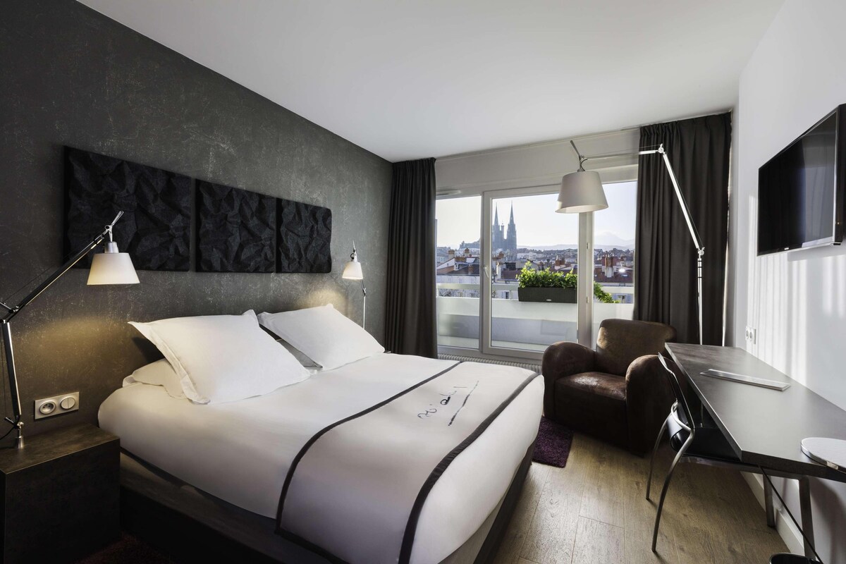 A modern hotel room with a large bed, a desk, and a view of a cityscape.