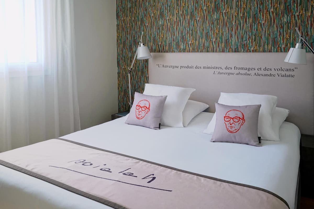 Una cama bien hecha con cabecera que incluye una cita, dos almohadas con diseño de rostro y una mesita de noche con lámpara.