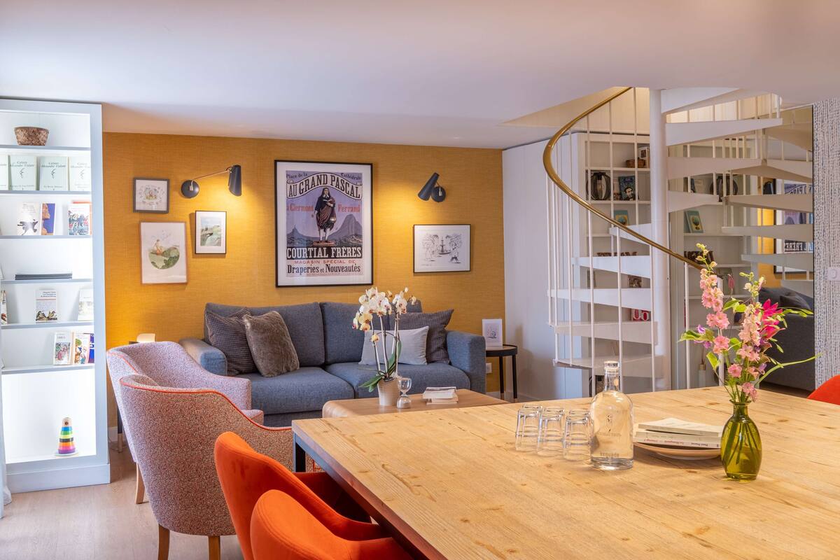 Una sala moderna con un sofá, una mesa de centro y sillas. Hay una estantería con libros y objetos decorativos. Un cartel y una obra de arte enmarcada decoran la pared.
