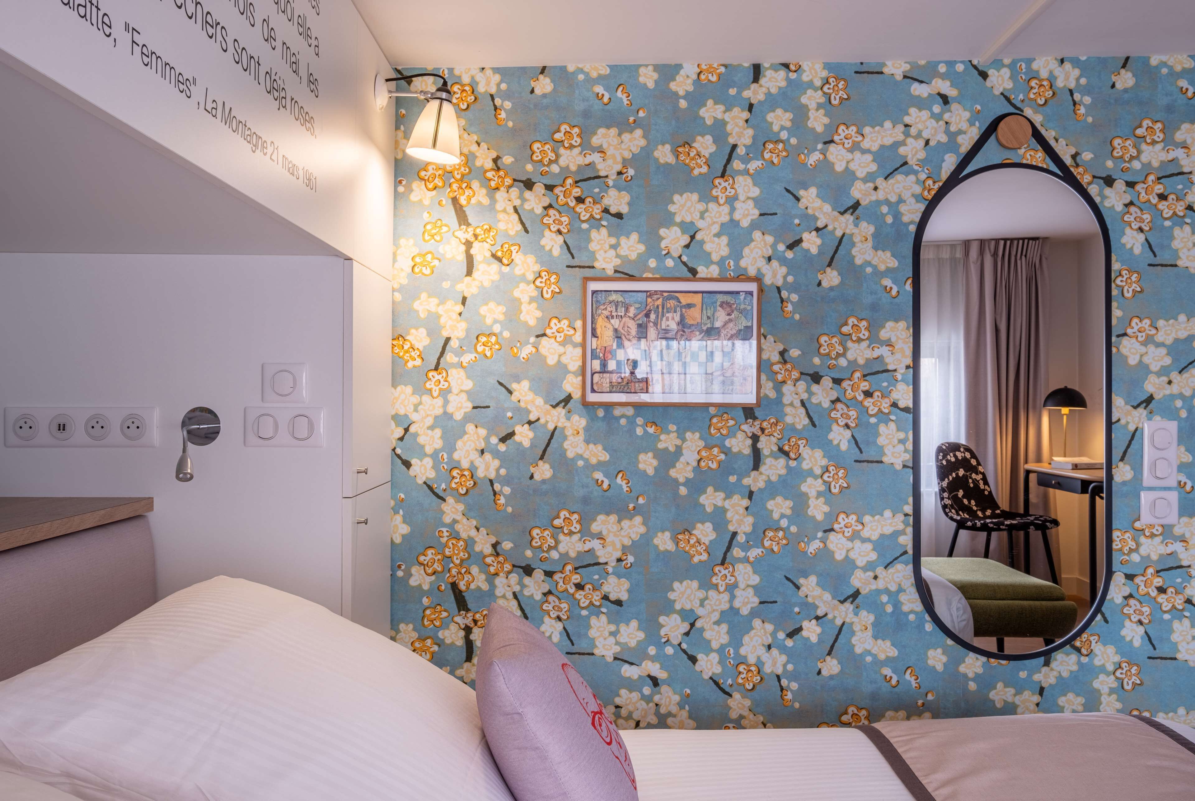 Une chambre d’hôtel avec un papier peint à motifs floraux, un lit avec des oreillers, une table de chevet, un miroir et un tableau encadré au mur.