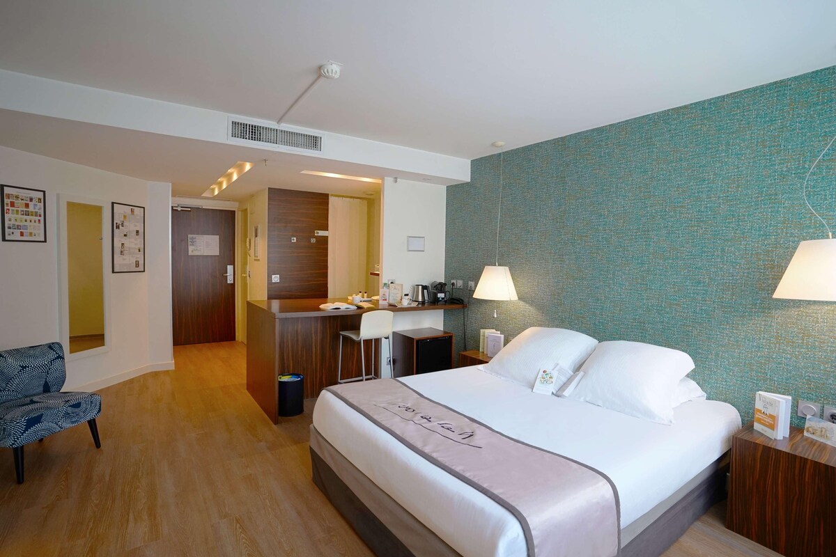 A hotel room with a large bed, a desk, a chair, and a small kitchenette.