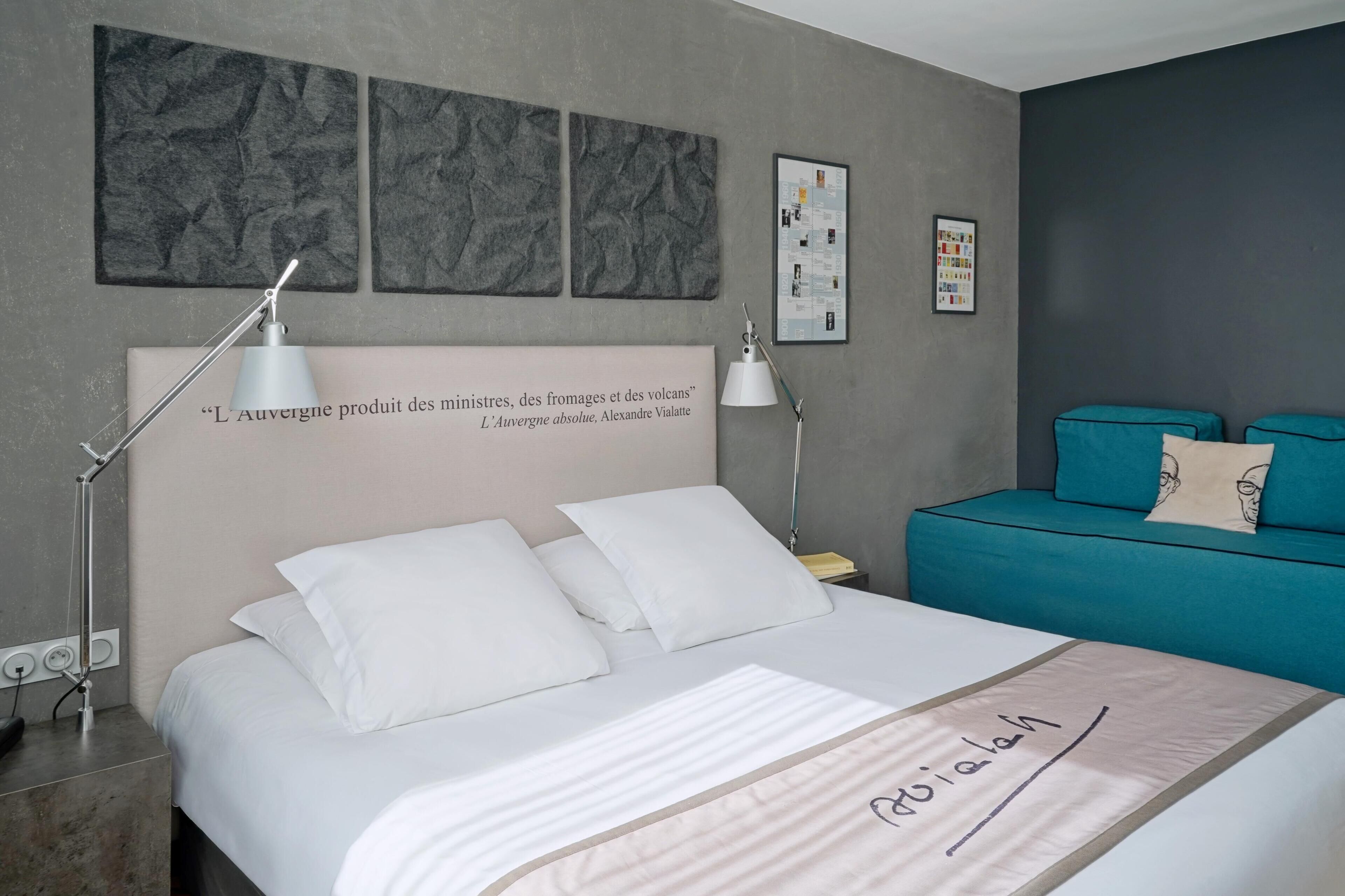 Moderni hotellihuone, jossa on siististi pedattu sänky, turkoosinsininen sohva ja taideteoksia seinillä.