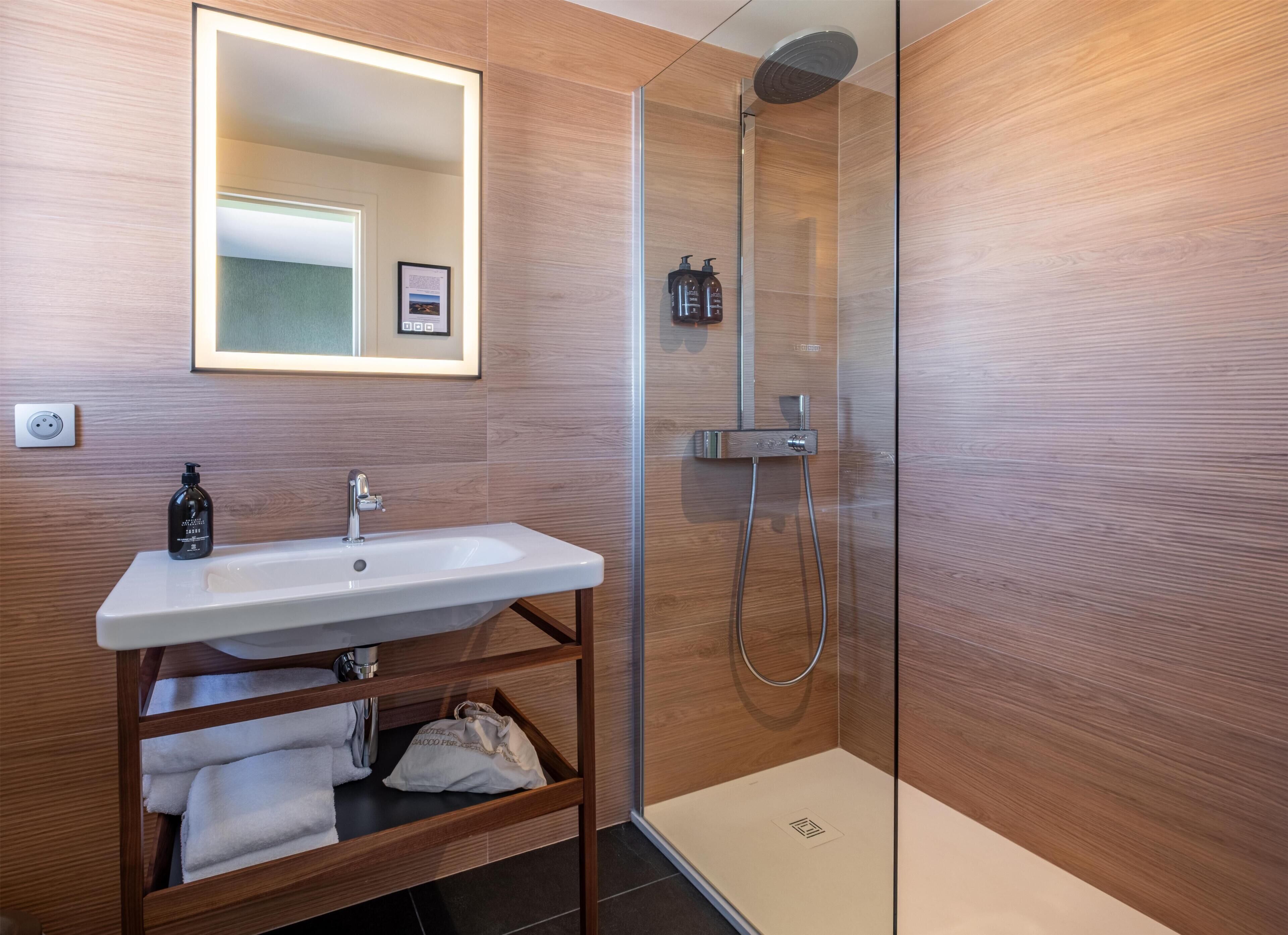 Un baño moderno con ducha envuelta en vidrio, un lavabo blanco y un mueble de tocador de madera.