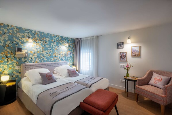 A hotel room with two beds, a red armchair, a small table, and a floral wallpaper.