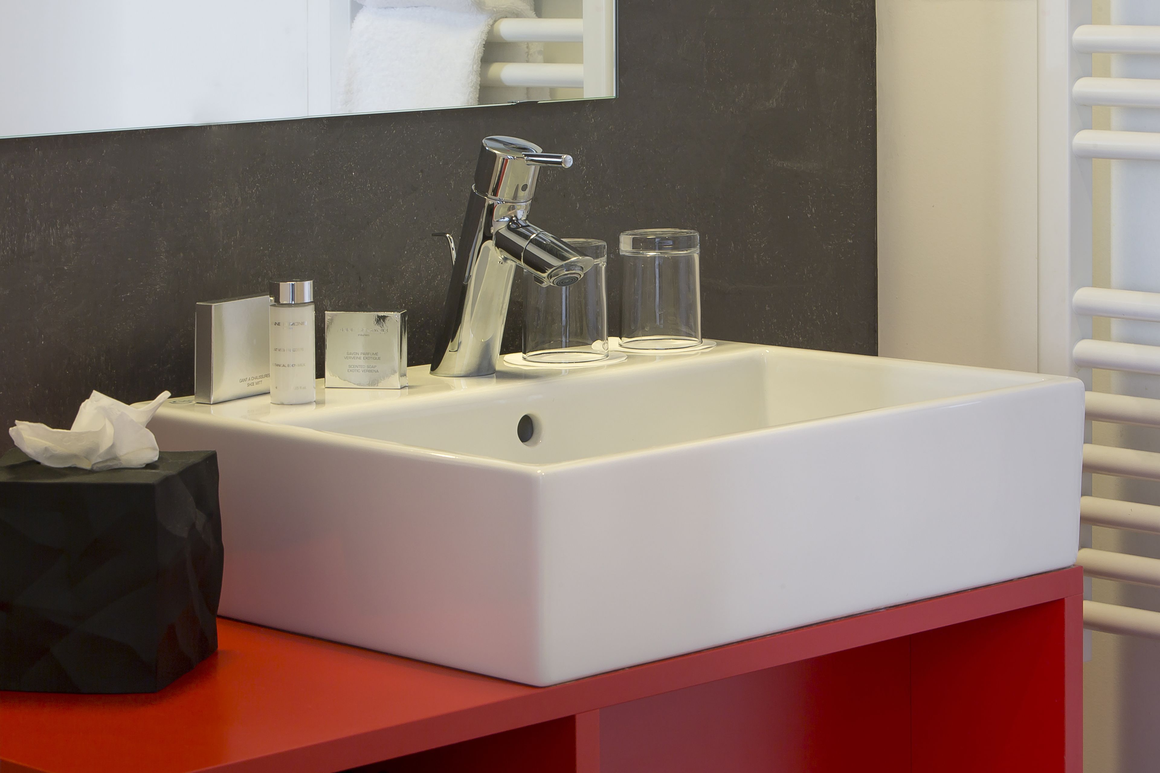 Une salle de bain moderne avec un lavabo blanc, un robinet chromé et un miroir au-dessus. On y trouve deux verres transparents sur le lavabo et une boîte à mouchoirs noire sur le comptoir.