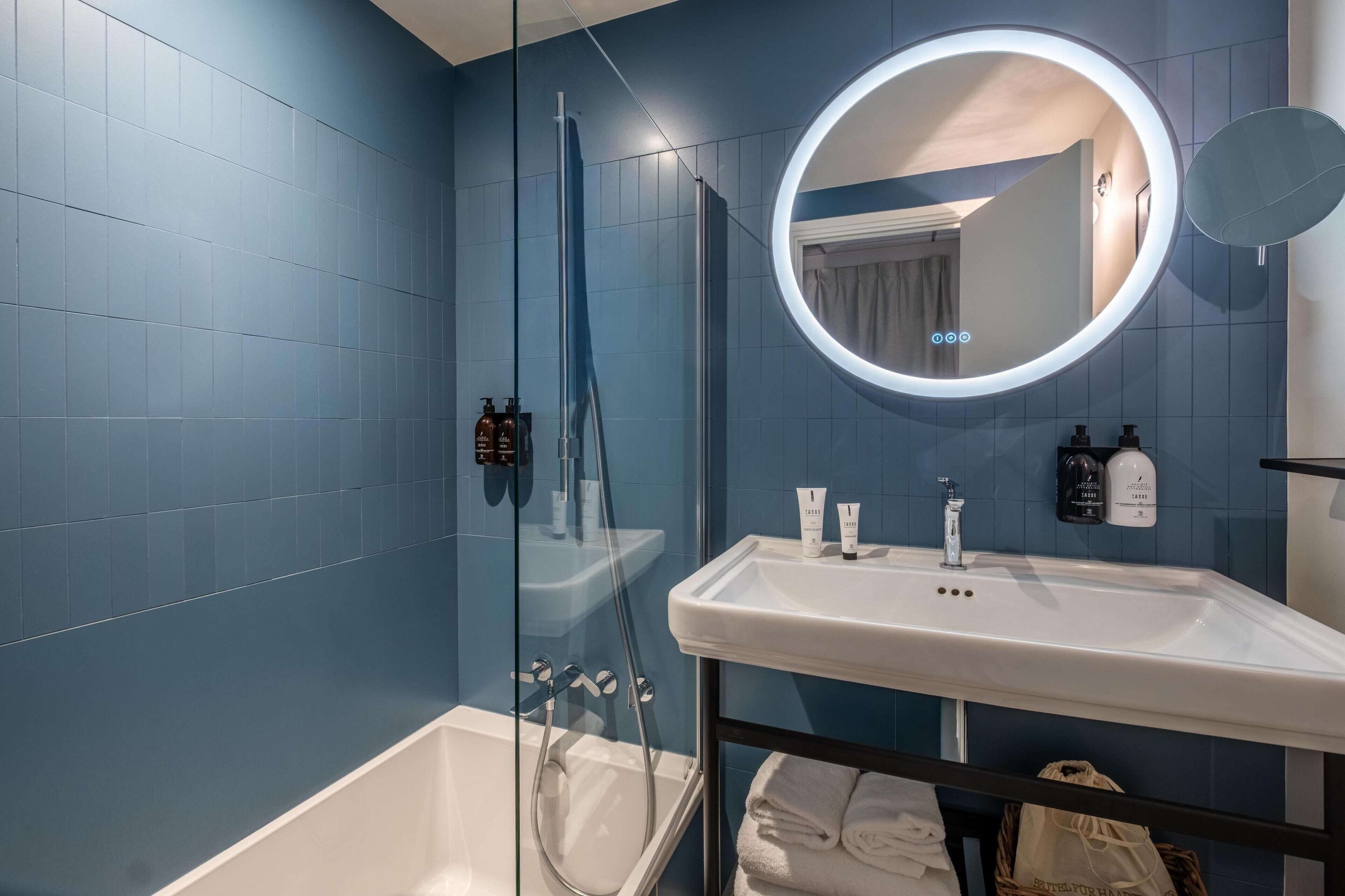 A modern bathroom with a glass-enclosed shower, a white sink, and a round mirror with a lighted frame.