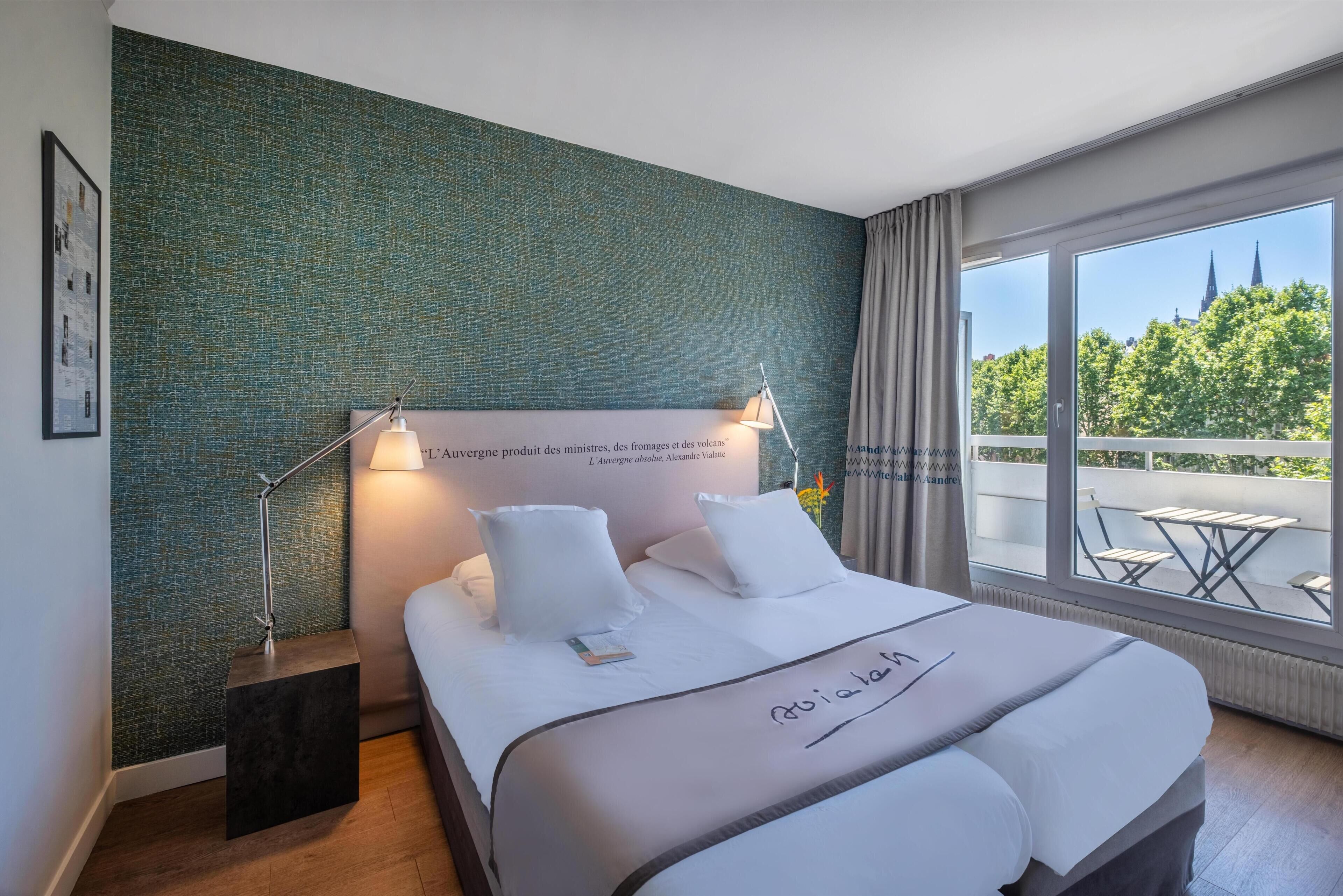 A hotel room with a large bed, a nightstand, a lamp, and a view of a cityscape through large windows.