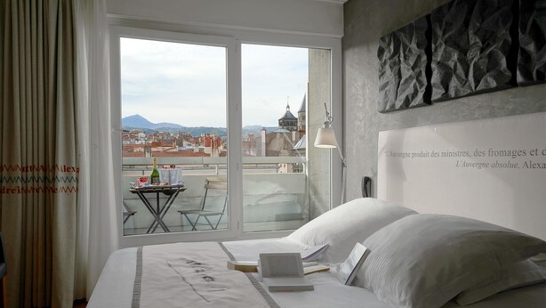 Una habitación de hotel con una cama, una computadora portátil y vistas a la ciudad a través de un ventanal amplio.