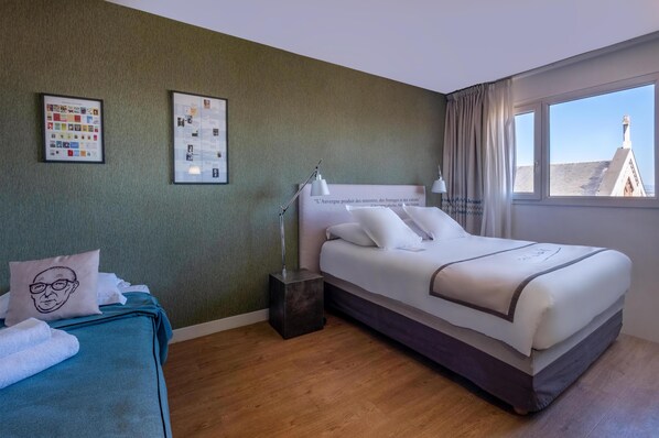 A hotel room with a bed, a bedside table with a lamp, a window with curtains, and two framed pictures on the wall.