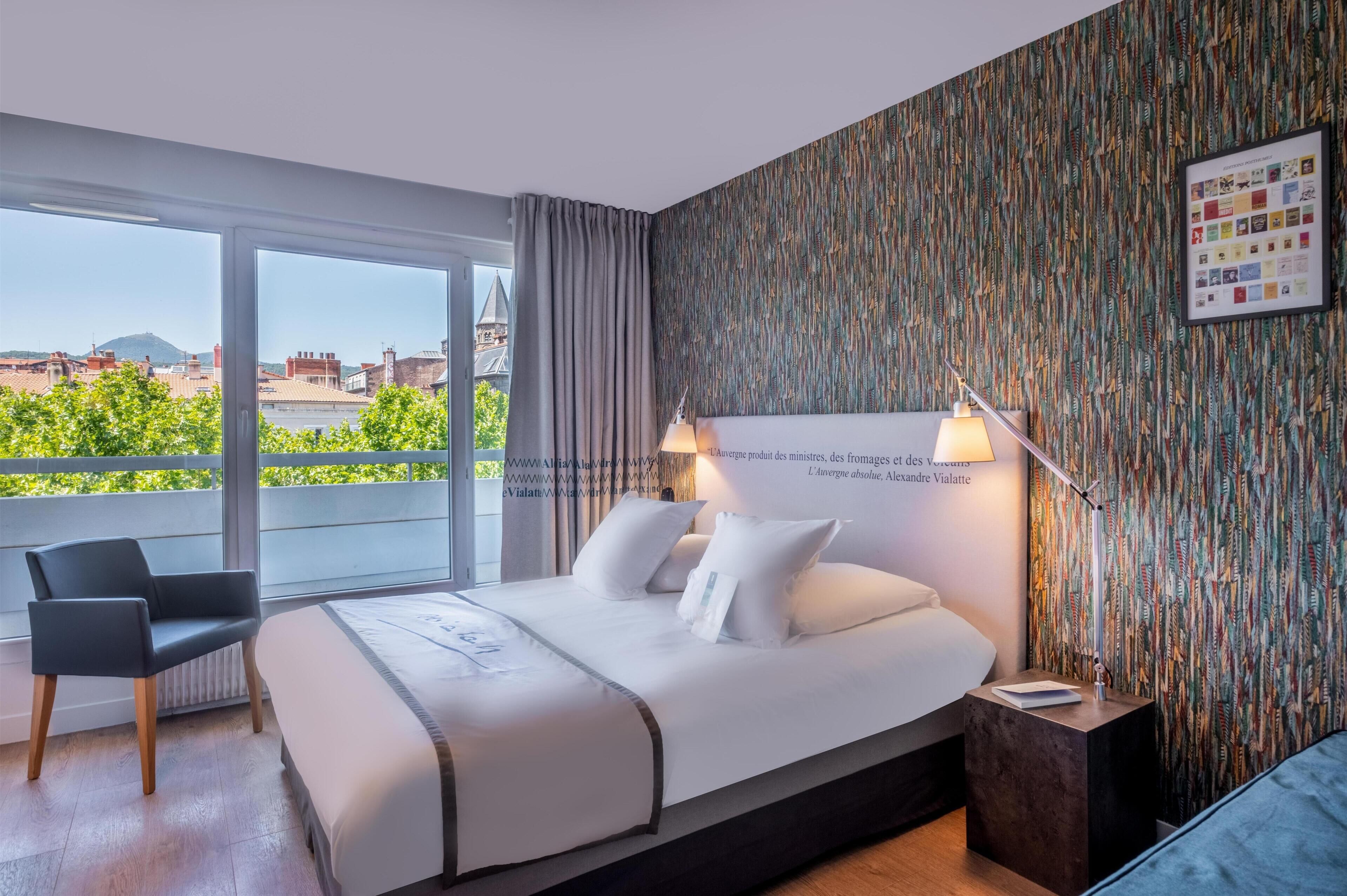 A hotel room with a large bed, a chair, a nightstand, and a view of buildings and greenery.
