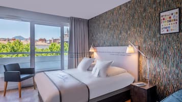 Quarto de hotel com uma cama grande, uma cadeira, uma mesa de cabeceira e vista para edifícios e vegetação.