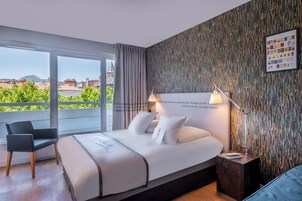 Habitación de hotel con una cama grande, una silla, una mesita de noche y vistas a edificios y zonas verdes.