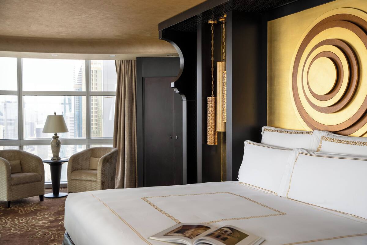 Habitación de hotel con una cama grande, dos sillones, una mesita de noche con lámpara y vistas a la ciudad a través de amplias ventanas.