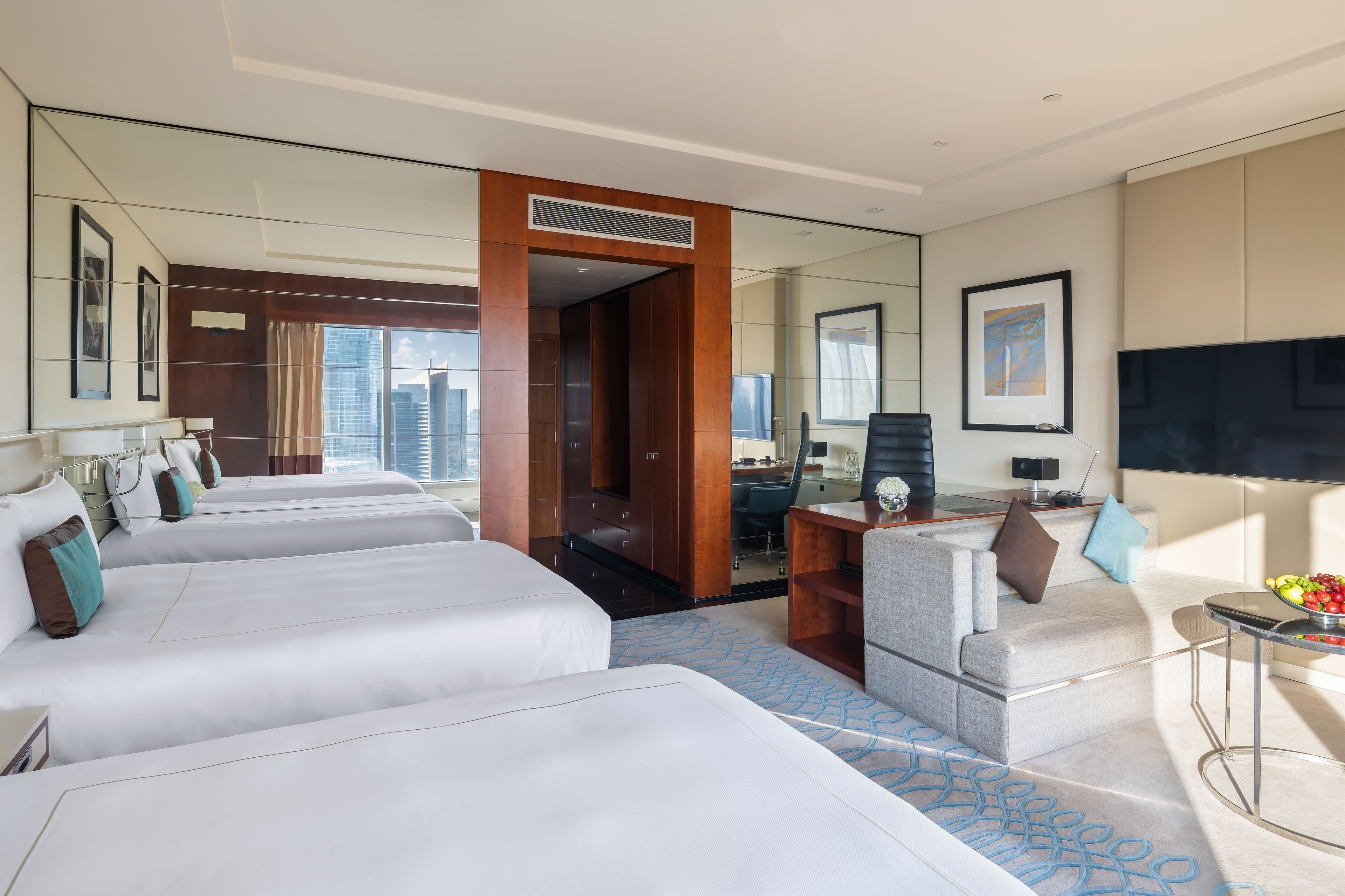 A hotel room with three beds, a desk, a sofa, a TV, and a large window with a city view.