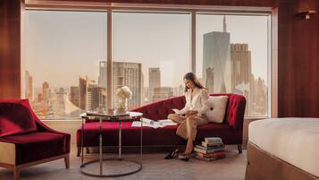 A modern hotel room with a large window offering a city view, a red sofa, a small round table with a vase, and a stack of books.