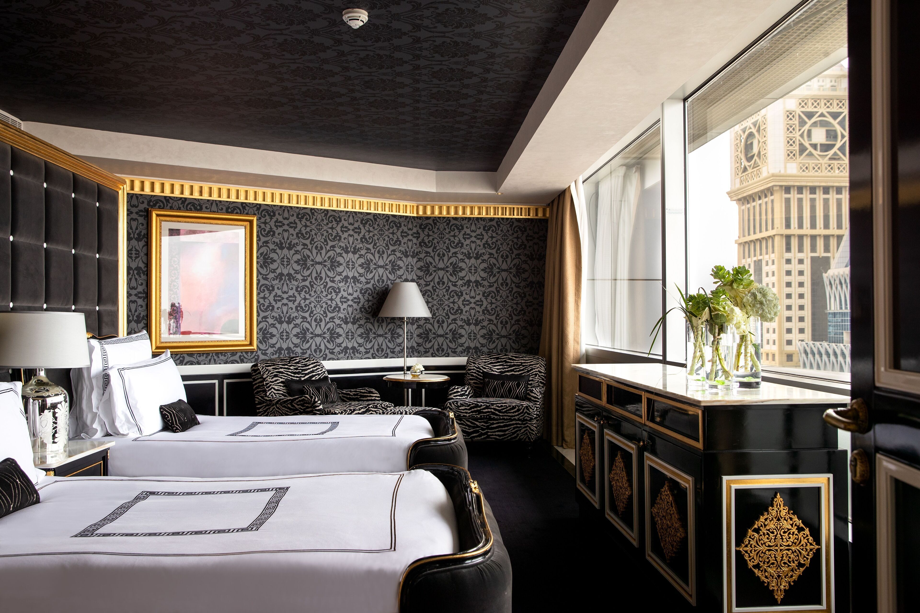 Ein Hotelzimmer mit zwei Betten, einem Schreibtisch, einem Stuhl, einem Fenster mit Blick auf ein Gebäude und einem dekorativen Spiegel.