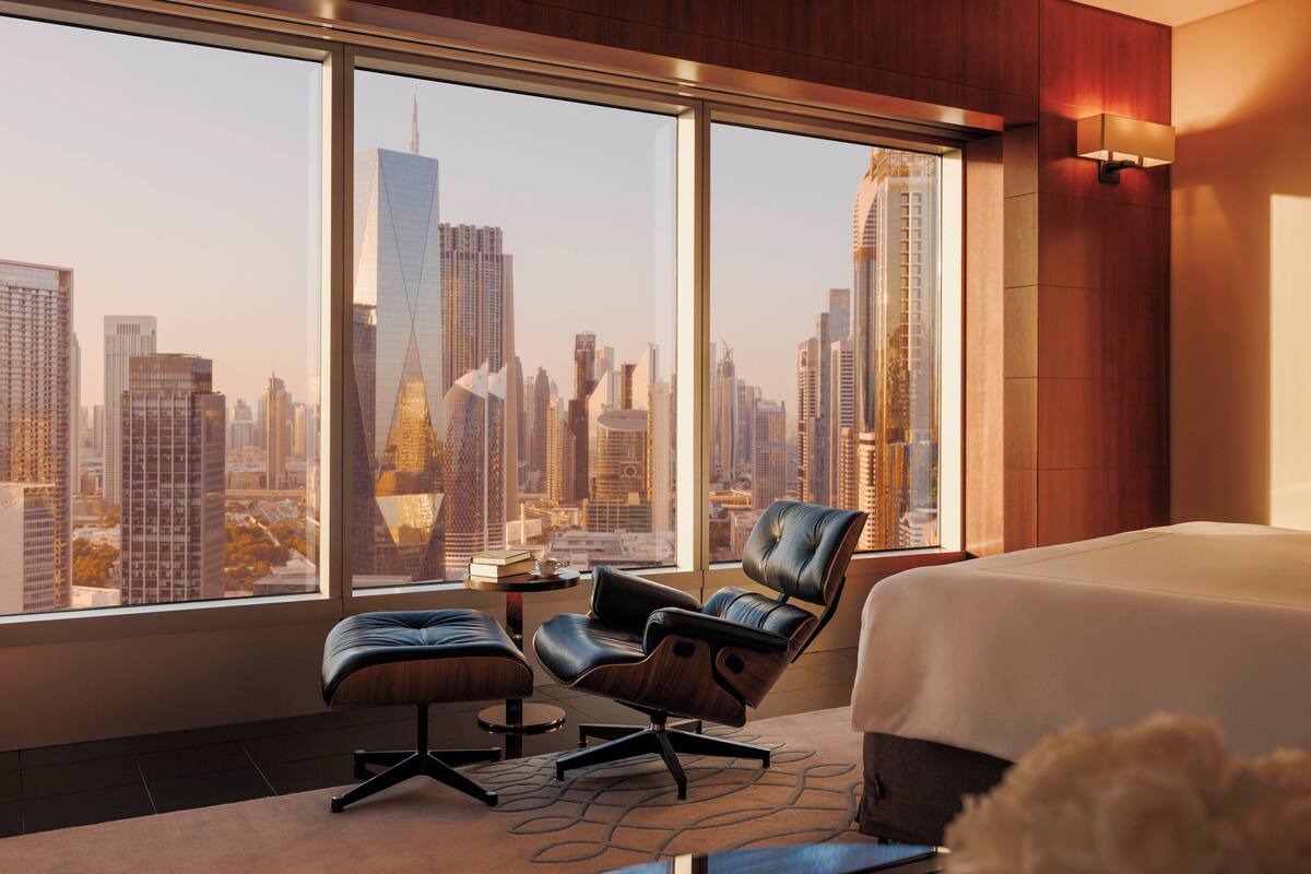 Habitación de hotel con un ventanal que ofrece vistas a un horizonte urbano, un sillón de cuero, una mesita con un libro y una cama con edredón blanco.