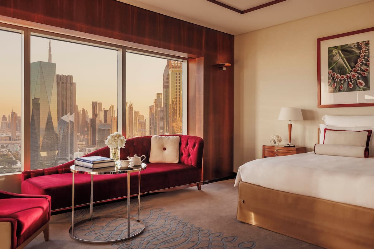 Habitación de hotel con un amplio ventanal que ofrece vistas del horizonte urbano, un sofá rojo, una cama y una pequeña mesa con un jarrón de flores.