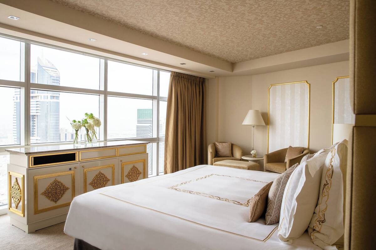 Habitación de hotel con una cama grande, un escritorio con lámpara, una silla y vistas a la ciudad a través de amplios ventanales.