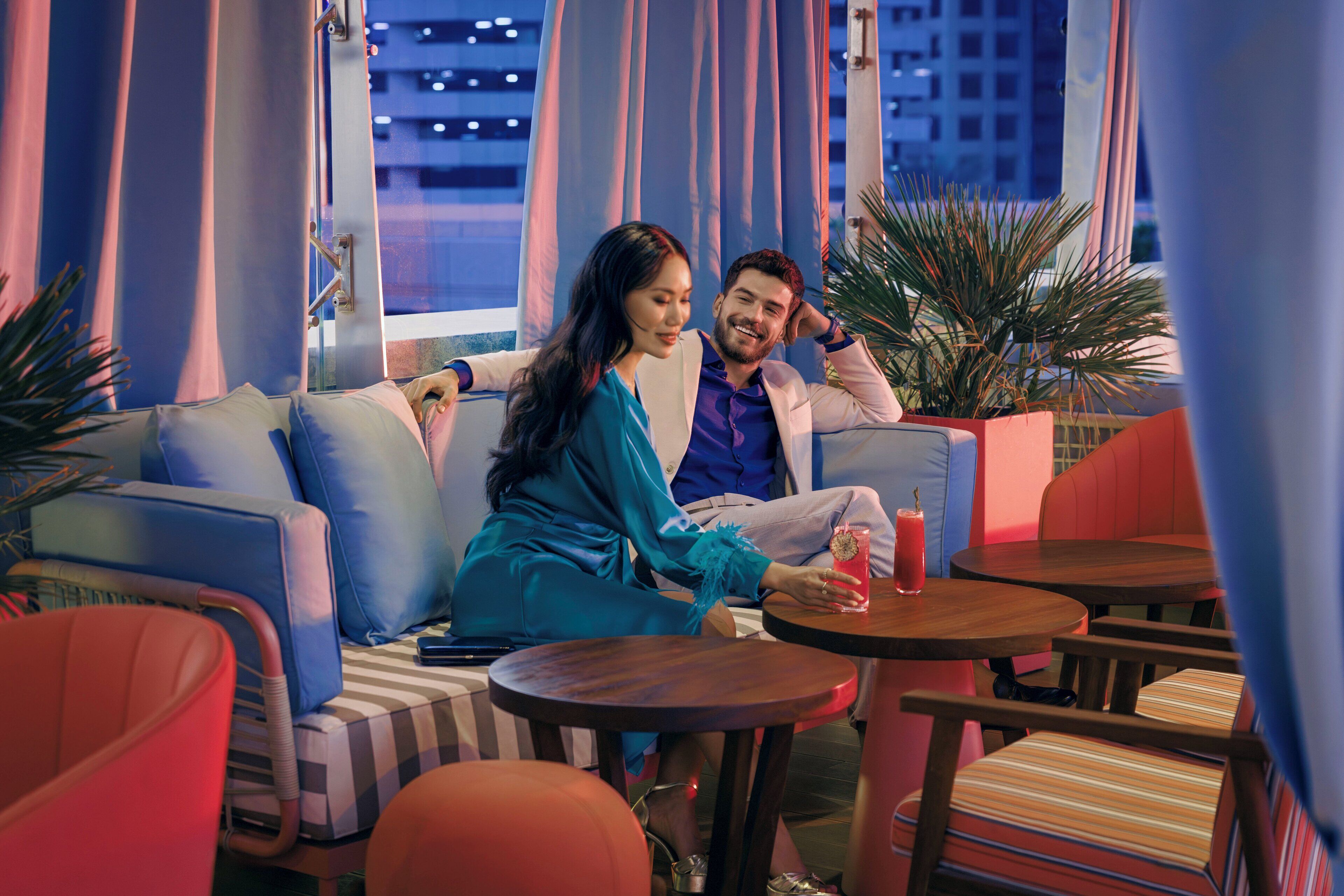 A couple sitting on a blue sofa in a modern lounge area with a city view, a small table with a drink, and red and orange chairs.