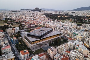 Un bâtiment moderne doté d’un toit plat et de grandes fenêtres, entouré d’un quartier urbain dense, avec divers immeubles et une montagne en arrière-plan.