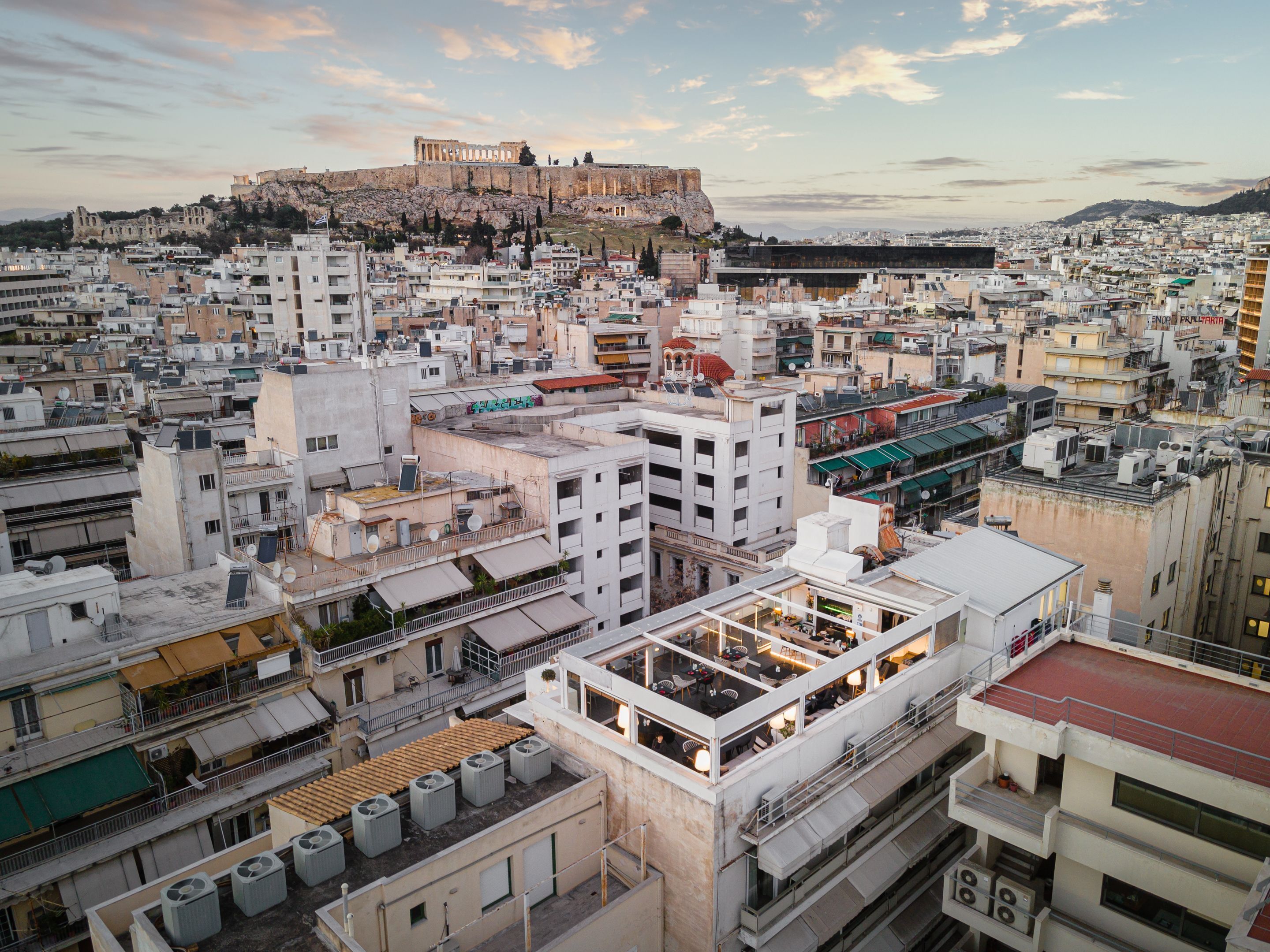A cityscape with a prominent ancient fortress on a hill, modern buildings, and a rooftop bar with outdoor seating.