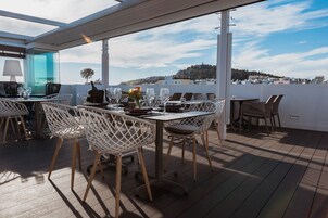 Terraço no telhado com mesa posta para refeições, cercado por cadeiras de vime, e vista para a paisagem urbana.