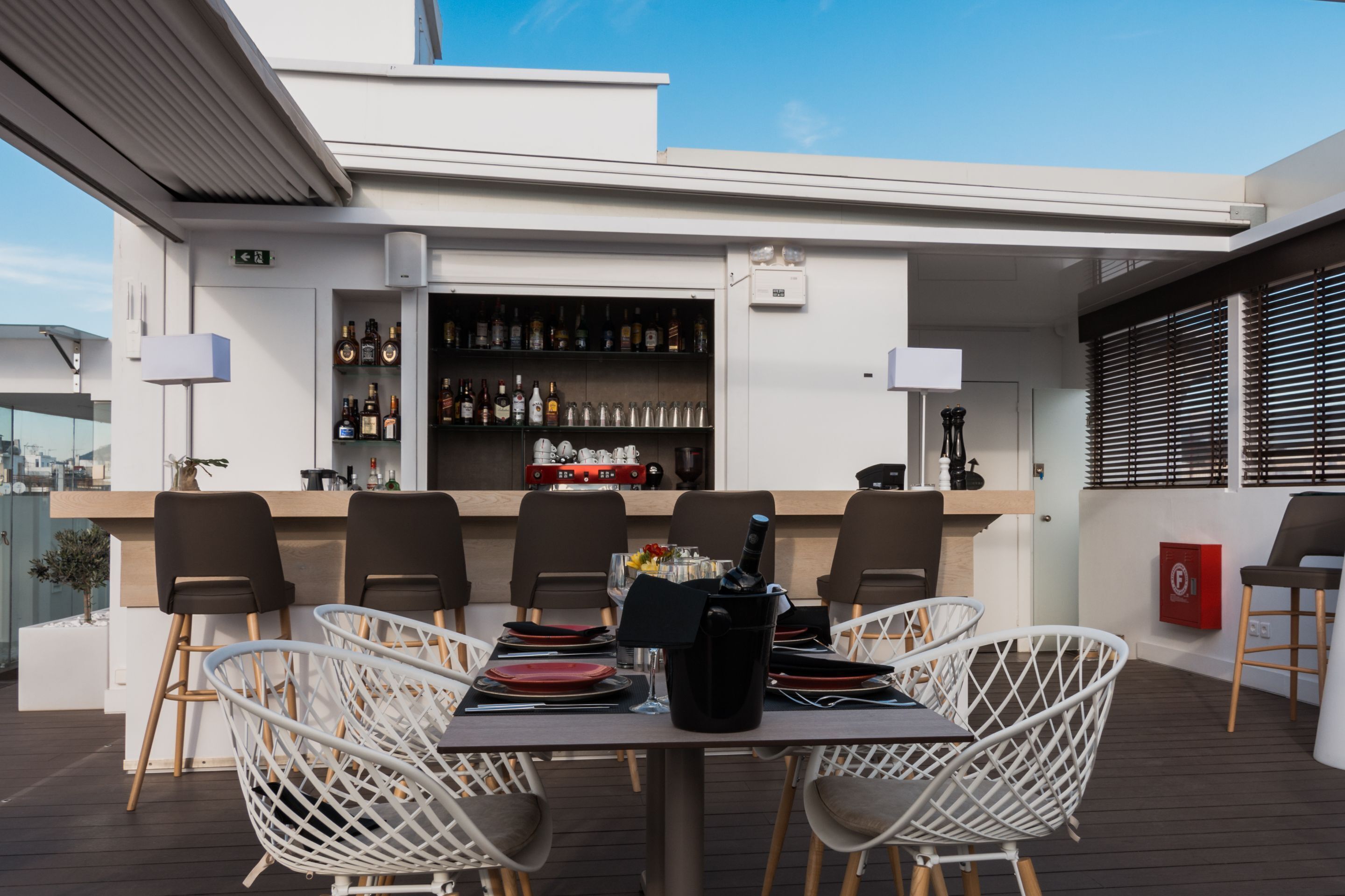 A rooftop bar with a bar counter, shelves of liquor bottles, white wicker chairs, and a table set for dining.