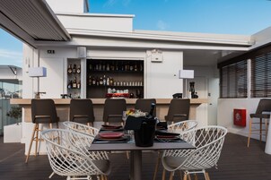 Um bar no terraço com balcão, prateleiras cheias de garrafas de bebidas, cadeiras de vime branco e uma mesa posta para refeições.
