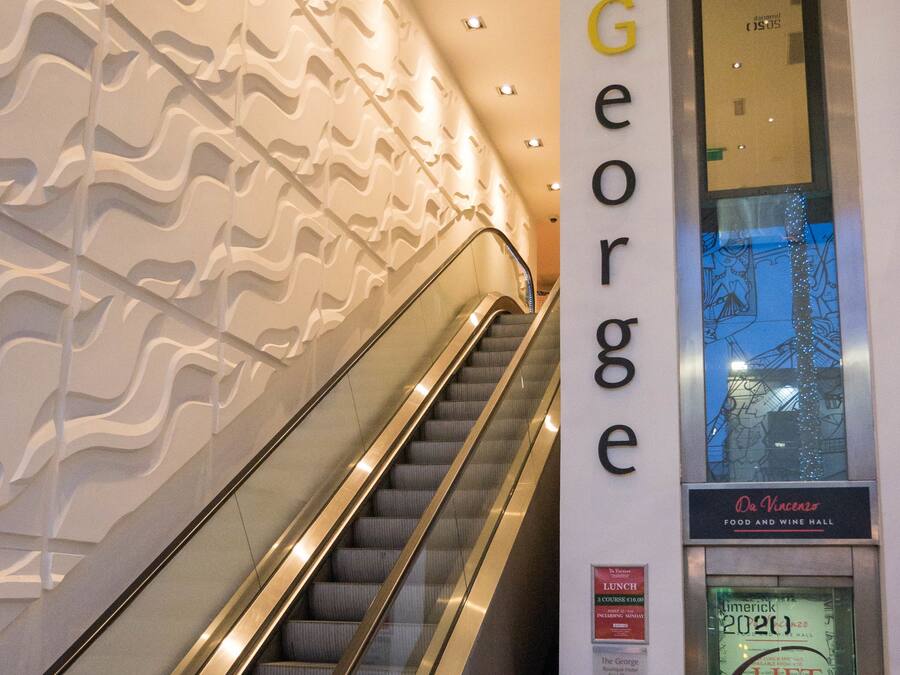 Una escalera mecánica en un edificio con un patrón ondulado en la pared y un letrero que indica el restaurante 'The George'.