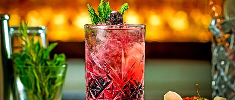 A bar setting with a glass of red cocktail garnished with mint, ice, and a black berry, accompanied by small bowls of appetizers and a glass of water.