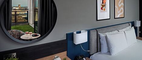 A modern hotel room with a round mirror, a bed with white linens, a bedside table with a vase and a phone, and a view of the city through the window.