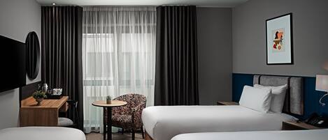 Habitación de hotel con dos camas, un escritorio con silla, una mesita pequeña y un ventanal grande con cortinas.