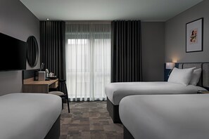 Ein Hotelzimmer mit zwei Betten, einem Schreibtisch mit Stuhl, einem Fernseher und einem Fenster mit Vorhängen.