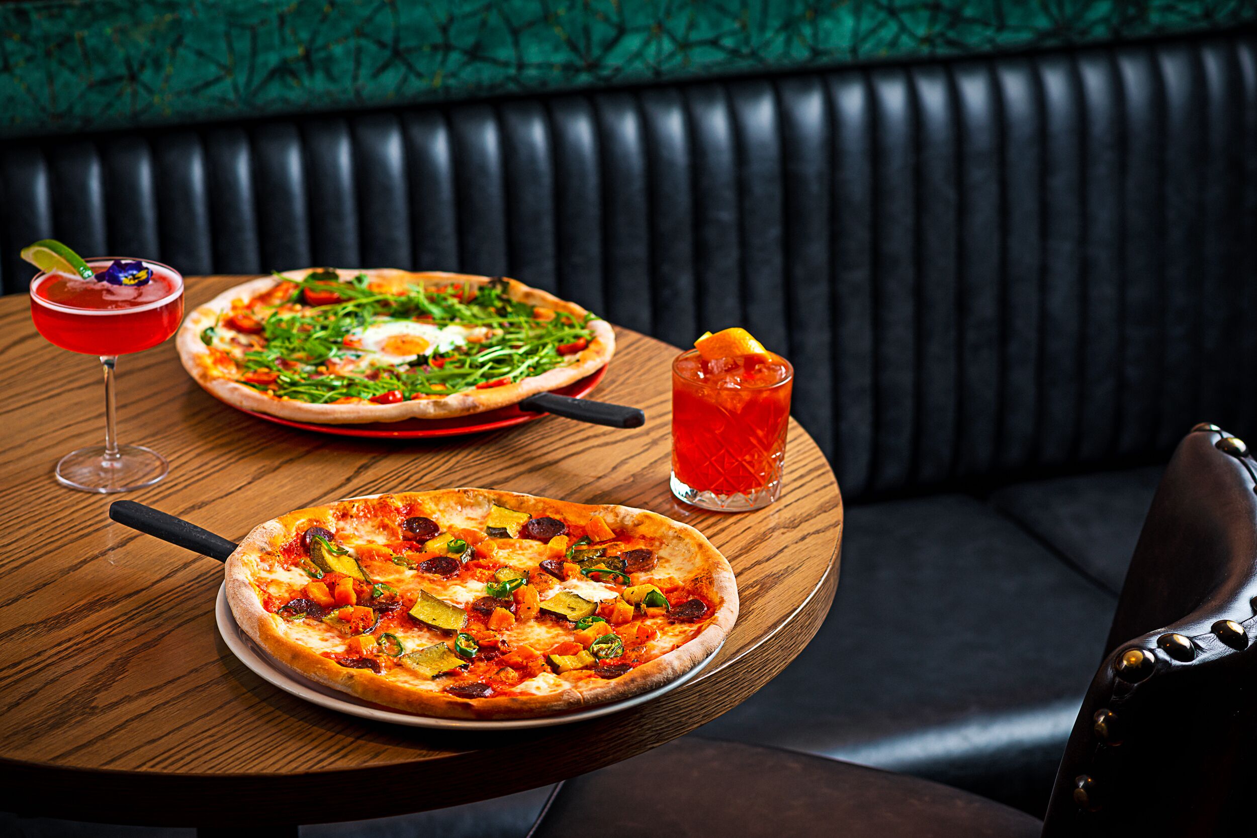 A pizza and a cocktail on a wooden table in a restaurant with a black leather booth in the background.