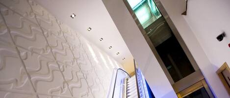 An escalator in a modern building with a wavy wall pattern and a glass elevator door in the background.
