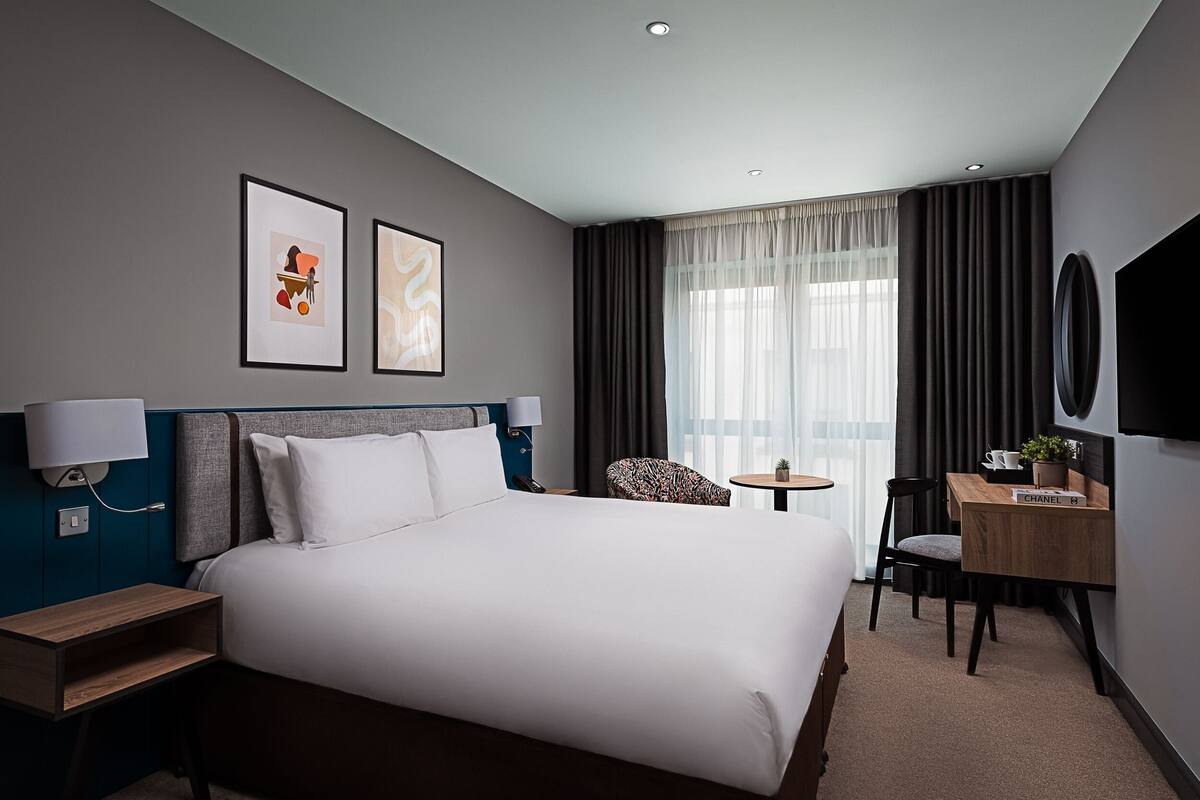 Quarto de hotel com uma cama grande, uma escrivaninha com cadeira, uma mesinha e duas quadros na parede.