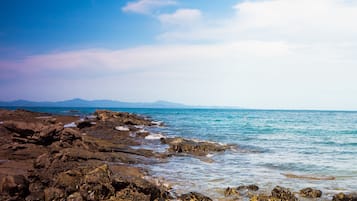 Una costa rocosa con aguas azules cristalinas y montañas distantes bajo un cielo parcialmente nublado.