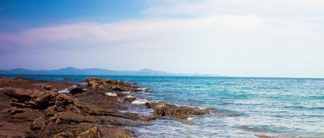 A rocky shoreline with clear blue water and distant mountains under a partly cloudy sky.