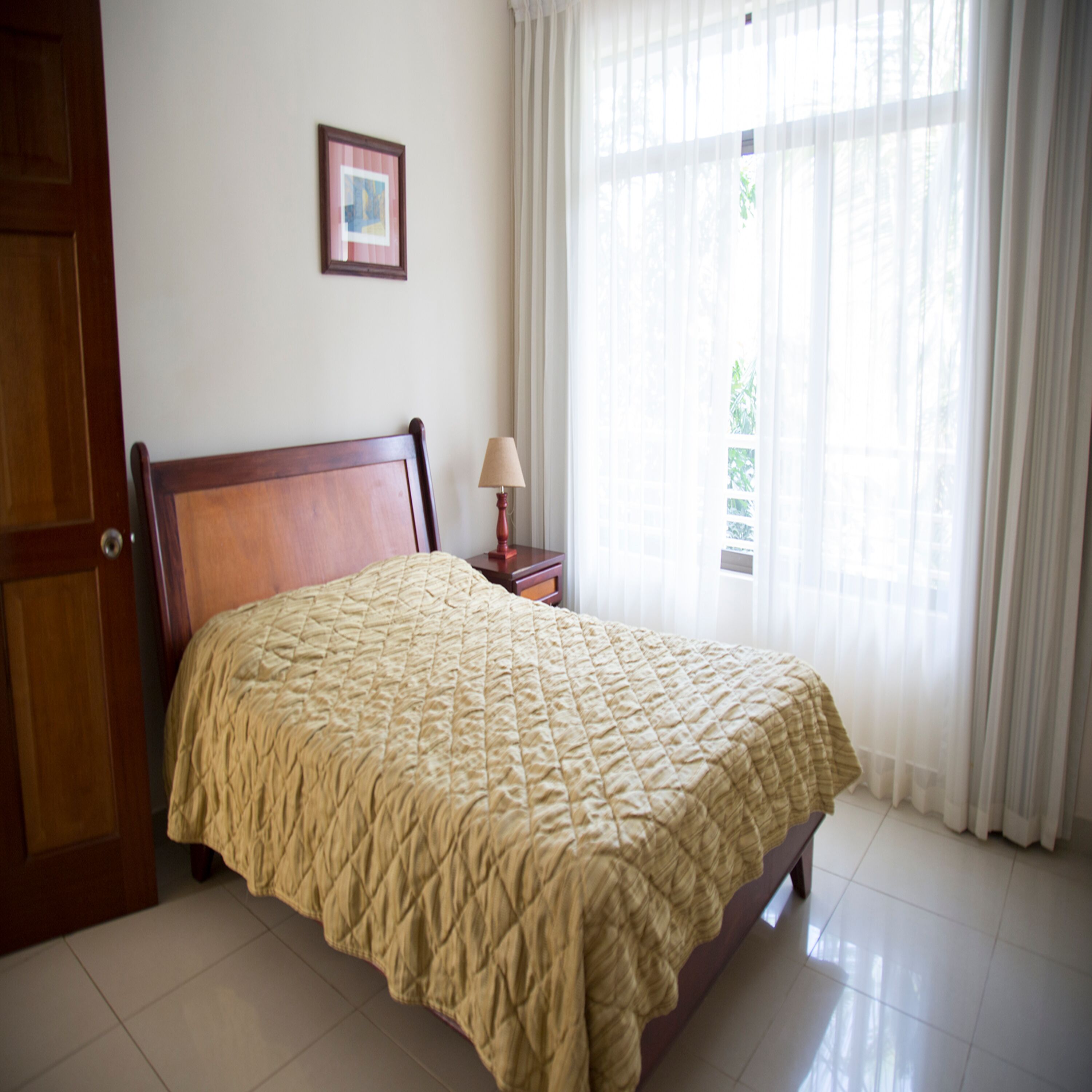 A bedroom with a wooden bed, a bedside table with a lamp, a framed picture on the wall, and sheer curtains on the window.