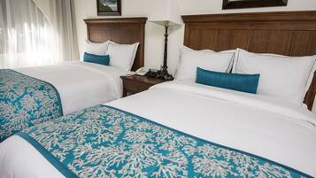 A hotel room with two beds, each with white bedding and blue decorative pillows. There is a wooden headboard, a bedside table with a lamp, and a framed picture on the wall.