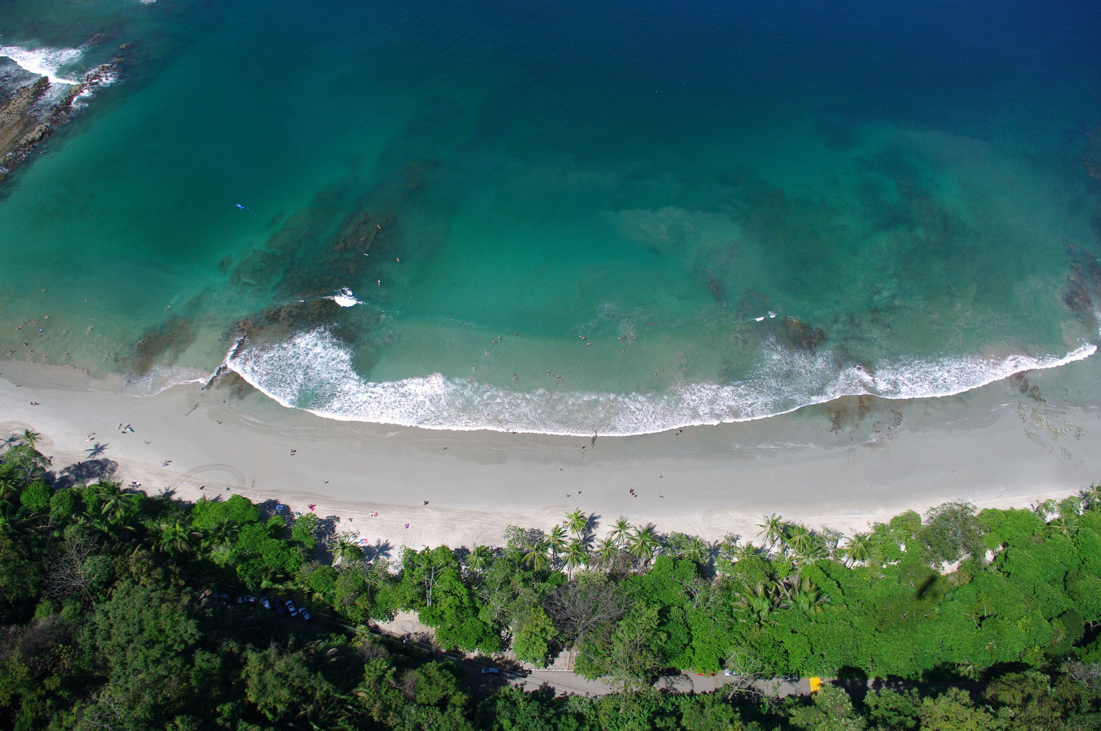 An aerial view of a beach with clear turquoise water, a sandy shore, and dense green vegetation at the beach's edge.