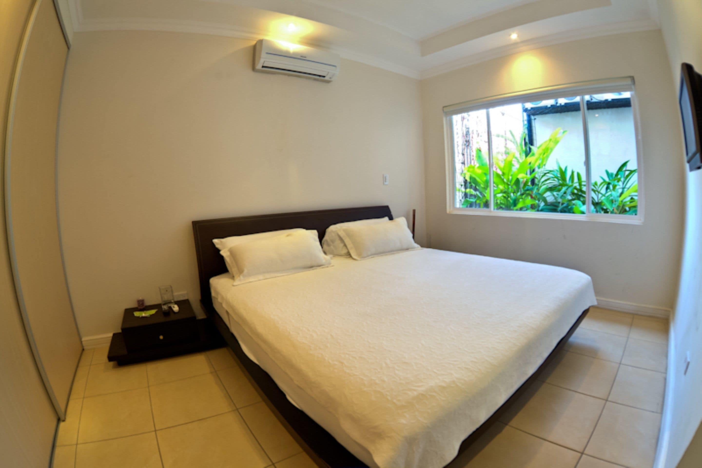 A bedroom with a large bed, a nightstand, an air conditioning unit, and a window with a view of greenery.