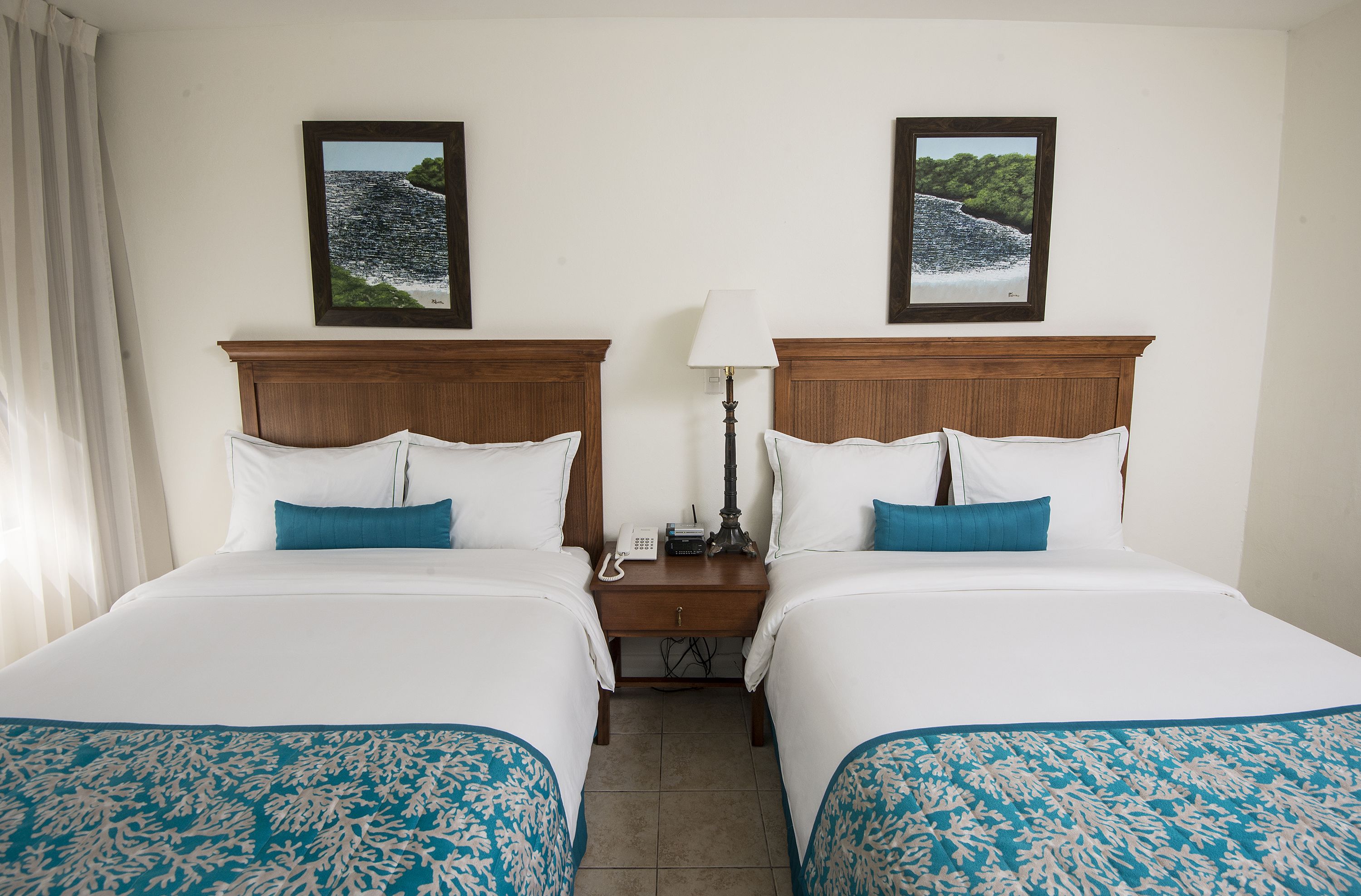 A hotel room with two beds, each with white bedding and blue accents, a nightstand with a lamp, and framed pictures on the wall.