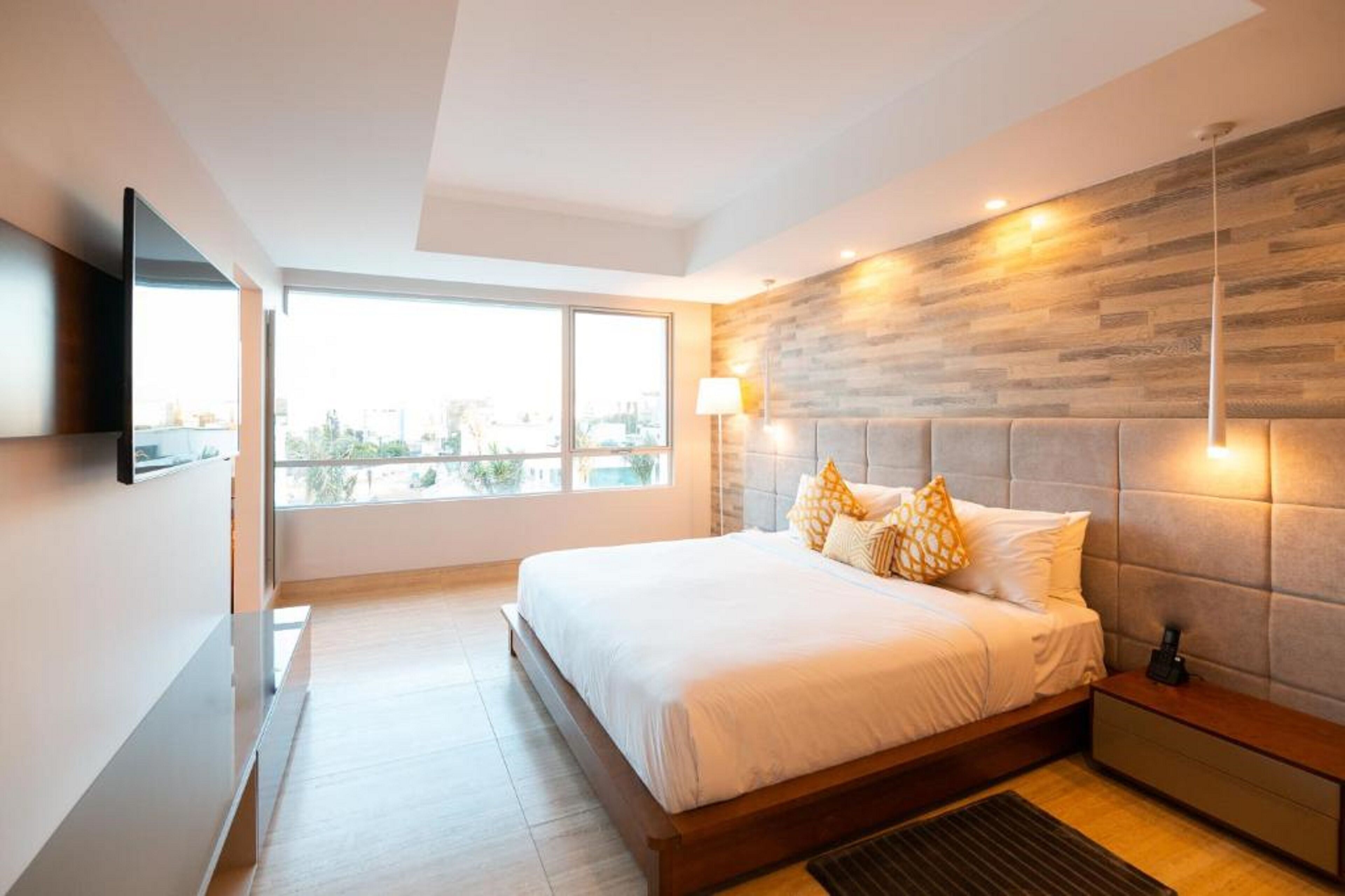 A modern bedroom with a large bed, a mounted TV, a city view, and a wooden accent wall.
