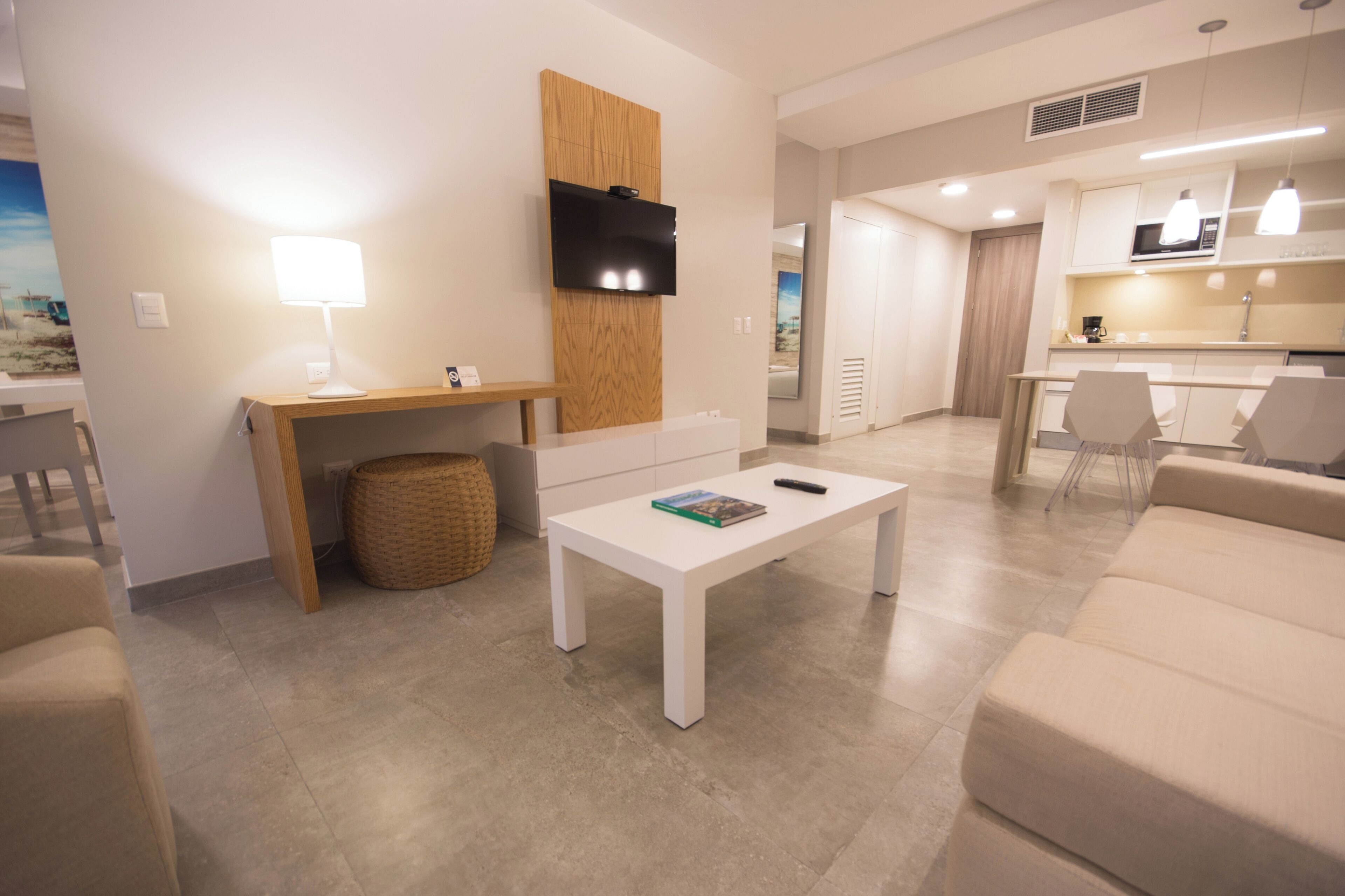 A modern living room with a white coffee table, a TV mounted on a wooden panel, and a kitchen area in the background.